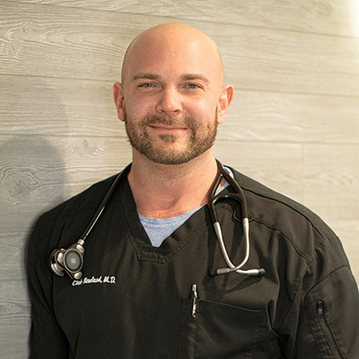 A man with a shaved head and beard stands against a light wood-paneled wall, wearing black medical scrubs with a stethoscope around his neck. The name “Chad Handschuh, M.D.” is embroidered on his uniform.