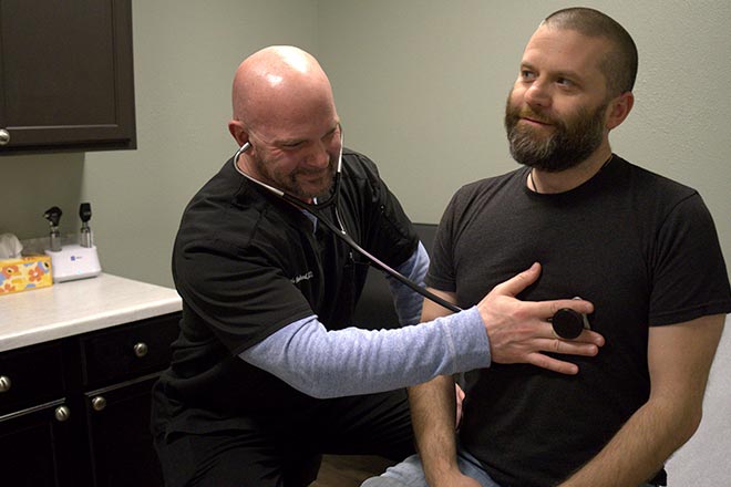 A healthcare professional uses a stethoscope to listen to the chest of a seated man during a medical examination in a clinic room.